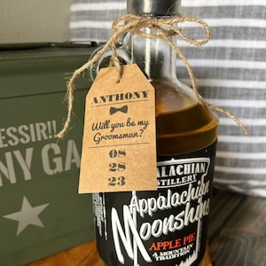 May include: A brown paper tag with the text "ANTHONY Will you be my Groomsman? 08 28 23" tied to a clear glass bottle with a black label that reads "Appalachian Stillery Moonshine Apple Pie A Mountain Tradition".