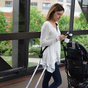 May include: A woman in a white sweater pushes a black stroller with a silver stroller handle extension. The stroller has a black and white cup holder.