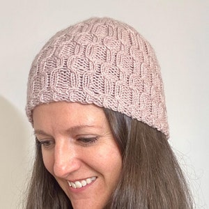 May include: A light pink knitted beanie hat with a textured, woven pattern. The hat is made of a soft, warm material and is designed to fit snugly on the head. This winter hat is perfect for keeping warm in cold weather.