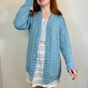 Pode incluir: Um cardigan de malha azul claro de manga comprida e decote em V. O cardigan é aberto na frente e tem uma bainha canelada.