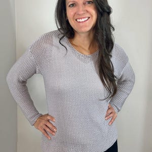 Puede incluir: Un suéter de punto gris claro con cuello en V. La parte superior de manga larga tiene un ajuste holgado y una apariencia texturizada. La mujer sonríe y lleva pantalones negros.