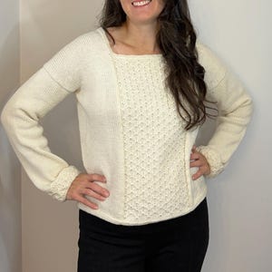 Puede incluir: Suéter de punto color crema con cuello cuadrado y mangas largas. El panel central presenta un patrón texturizado, mientras que las mangas y los lados tienen un tejido más simple. Los puños y el escote tienen un ribete decorativo. La persona lleva pantalones negros.