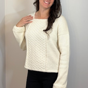 May include: Cream-colored sweater with a square neckline and textured knit panel. The long-sleeved sweater has ribbed cuffs and a relaxed fit. The woman in the image is wearing black pants.