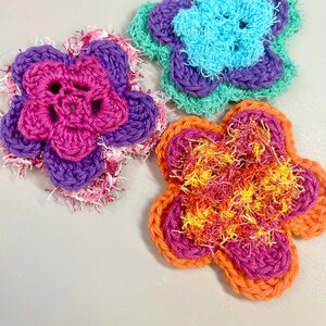 May include: Three crocheted flower shapes in different colors. The first flower is pink and purple with a white fringe. The second flower is turquoise with a purple and green outer layer. The third flower is orange with a pink and yellow inner layer.