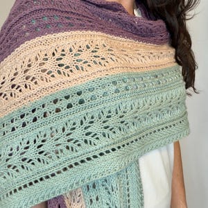 May include: A knitted shawl in shades of purple, beige, and mint green. The shawl features an openwork design with a striped pattern. The garment is draped over the shoulders.
