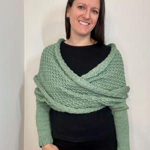 Puede incluir: Una envoltura de punto verde salvia con mangas largas. La envoltura se lleva sobre una blusa negra y pantalones negros.