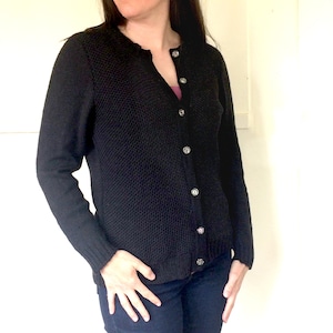 May include: A black knitted cardigan with a button-up front and long sleeves. The cardigan is worn over a pink shirt and blue jeans.