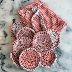 May include: A set of handmade crochet face scrubbies and a matching pouch. The scrubbies are circular, with a spiral design in pink and gray. The pouch is a solid pink with a drawstring closure. The items are arranged on a marble surface.