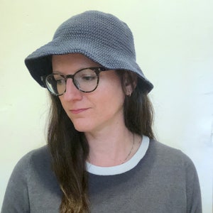 May include: A person wearing a dark blue bucket hat with a wide brim and black framed glasses. They are wearing a dark grey shirt with a white trim around the neck.