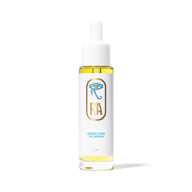 May include: A white bottle with a dropper top, filled with yellow oil. The label features a blue eye symbol and the gold letters "RA". Below, the text reads "Desert Date Oil Serum" and "30 mL".