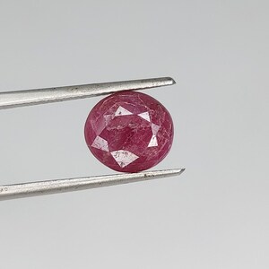 Pode incluir: Uma gema de rubi redonda e facetada, segurada por pinças prateadas. A gema é de cor vermelha profunda e tem uma superfície polida e reflexiva. As pinças estão posicionadas contra um fundo cinza claro liso.
