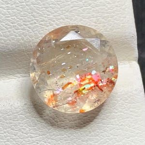 Puede incluir: Una piedra preciosa de piedra de sol redonda y facetada con una base beige pálido translúcida e inclusiones internas que muestran motas vibrantes, brillantes, naranjas, rojas e iridiscentes en toda la estructura.
