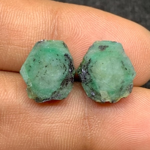 Könnte beinhalten: Zwei rohe Smaragd-Edelsteine. Die Smaragde sind leuchtend grün mit dunklen Einschlüssen. Die Steine haben eine sechseckige Form. Die Edelsteine werden vor einem neutralen Hintergrund gezeigt.