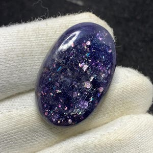 May include: An oval, dark blue gemstone with embedded iridescent glitter. The polished stone has a smooth surface and is held in white fabric. The glitter reflects pink, purple, and blue hues.