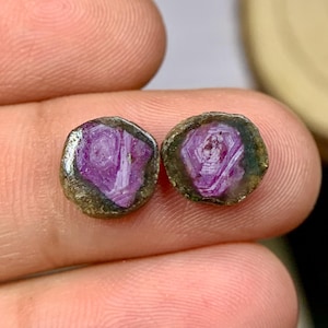 Puede incluir: Dos gemas en bruto, sin cortar, con un interior púrpura intenso y un borde exterior verde-marrón oscuro. Las gemas son redondas y parecen estar sostenidas en una mano. El color púrpura es vibrante y el borde exterior tiene una textura rugosa.