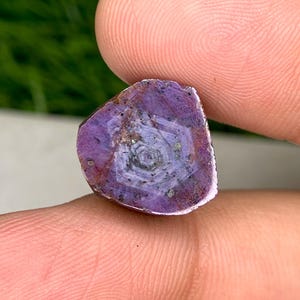 May include: A close-up of a raw, uncut gemstone. The stone is a deep purple colour with a star-like pattern in the centre. The edges of the stone are rough and uneven. Small flecks of black are scattered throughout the stone.