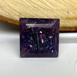 May include: A square-cut, deep purple gemstone with internal iridescent inclusions. The gem has a polished surface. The gemstone's facets catch the light, creating a sparkling effect. The gem is set against a light background.