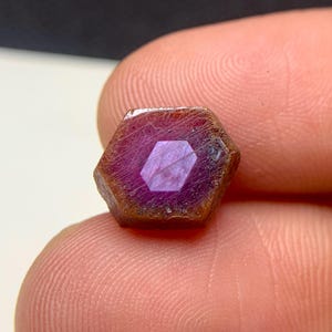 Könnte beinhalten: Ein facettierter, sechseckiger Rubin-Edelstein mit tiefmagentafarbener Tönung. Der Edelstein hat eine polierte Mitte und eine raue, braune Außenseite. Der Rubin wird zwischen zwei Fingern gehalten, um seine Größe und Details zu zeigen.