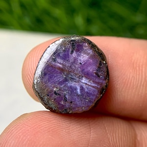 Könnte beinhalten: Ein polierter, oval geformter Edelstein mit tiefvioletter Farbe und einem sternartigen Muster. Der Stein hat einen dunklen, rauen äußeren Rand und eine glatte, reflektierende Oberfläche. Der Sterneffekt wird durch sich kreuzende Linien erzeugt.