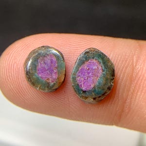 May include: Two oval gemstones with a vivid purple core, framed by a dark green and brown border. The close-up view highlights the natural textures and colours of the stones. These gemstones are likely intended for jewellery.