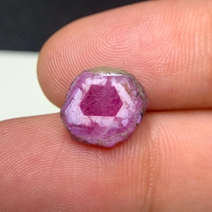 Puede incluir: Una gema en bruto sin cortar con un interior hexagonal. La gema es de color rosa intenso con un exterior rosa claro y gris. La piedra se sostiene entre dos dedos.