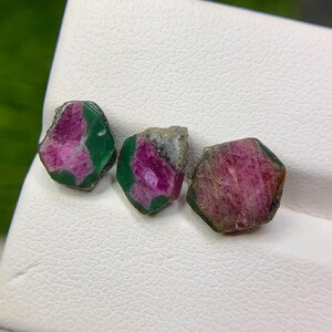 以下が含まれることがあります： 深紅と緑色が混ざった3つのラフカットルビーの原石。未研磨の石は白い表面に置かれ、自然な外観を際立たせています。宝石は形や大きさがわずかに異なります。