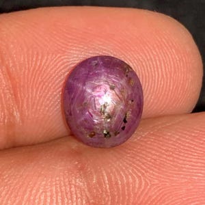 May include: A close-up of an oval-shaped, purplish-red gemstone. The gem has a polished surface with internal inclusions and a subtle play of light. The stone is held between fingers, showcasing its size and color.