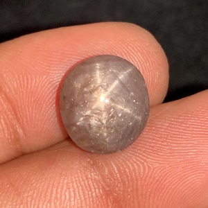 Puede incluir: Una gema redonda y gris con un patrón en forma de estrella en su superficie. La gema se sostiene en una mano, mostrando sus cualidades pulidas y reflectantes. El efecto de estrella se crea por la reflexión de la luz.