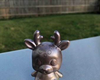May include: A small, silver-coloured deer figurine with large eyes and small antlers. The deer has a rounded body and is sitting upright. The figurine is made of a metallic material and has a smooth finish. The background is a blurred view of a garden.