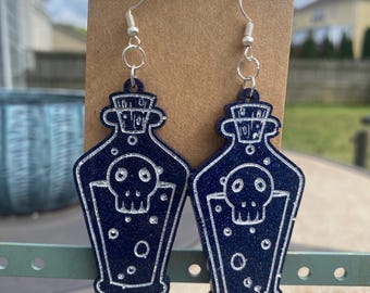 Peut inclure: Une paire de boucles d'oreilles en forme de flacons de potion bleu foncé avec un motif de tête de mort. Les boucles d'oreilles ont des crochets argentés et sont présentées sur une carte marron. Un accessoire effrayant.