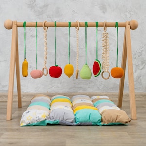 May include: Wooden baby play gym with hanging toys, including a banana, apple, pear, lemon, watermelon, and a ring. The gym is set up on a colorful patchwork play mat.