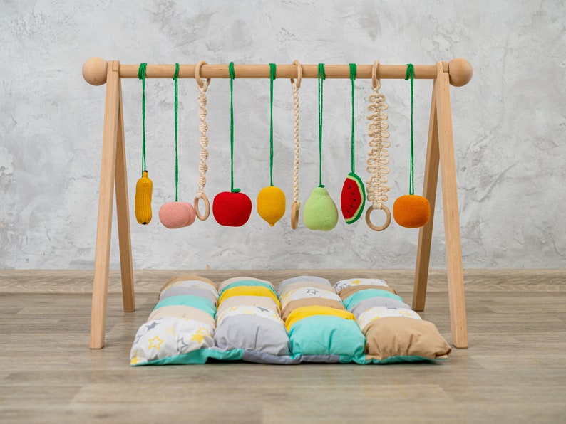 May include: Wooden baby play gym with hanging toys, including a banana, apple, pear, lemon, watermelon, and a ring. The gym is set up on a colorful patchwork play mat.
