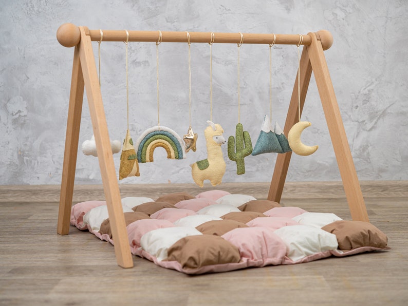 May include: A wooden play gym with a padded play mat. The gym has a wooden frame with a hanging bar and various soft toys, including a rainbow, a star, a cactus, a mountain, a cloud, and a llama.