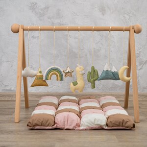 May include: A wooden play gym with hanging felt toys, including a cloud, mountain, rainbow, star, llama, cactus, mountains, and a crescent moon. The gym is set up on a brown and pink quilted play mat.