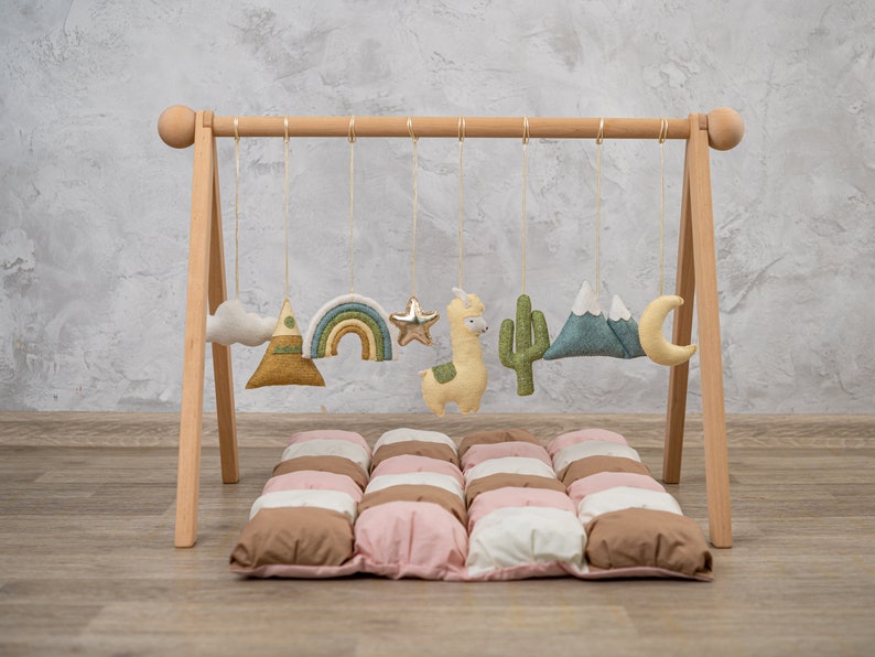 May include: A wooden play gym with hanging felt toys, including a cloud, mountain, rainbow, star, llama, cactus, mountains, and a crescent moon. The gym is set up on a brown and pink quilted play mat.