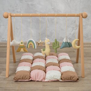May include: A wooden play gym with hanging toys for babies. The toys include a cloud, a rainbow, a star, a llama, a cactus, a mountain, and a moon. The play gym is set up on a padded play mat with brown, pink, and white squares.