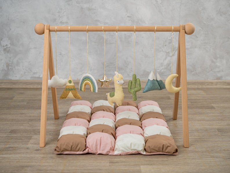 May include: A wooden play gym with hanging toys for babies. The toys include a cloud, a rainbow, a star, a llama, a cactus, a mountain, and a moon. The play gym is set up on a padded play mat with brown, pink, and white squares.