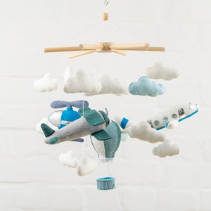 May include: A baby mobile with a blue and green airplane, a hot air balloon, and white clouds. The mobile is made of felt and is suspended from a wooden frame.