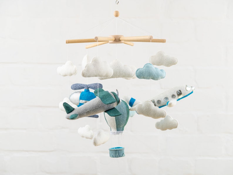 May include: A baby mobile with a blue and green airplane, a hot air balloon, and white clouds. The mobile is made of felt and is suspended from a wooden frame.