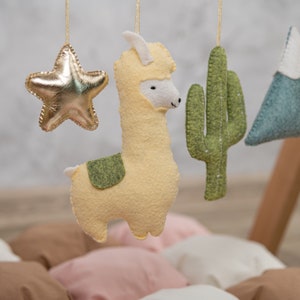 May include: A baby mobile with a yellow llama, a gold star, a green cactus, and a blue mountain range. The mobile is hanging from a wooden frame.