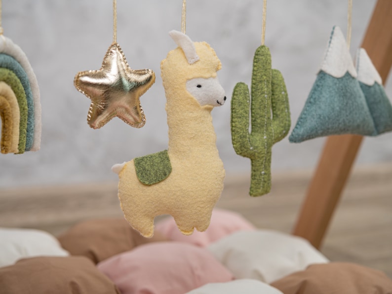 May include: A baby mobile with a yellow llama, a gold star, a green cactus, and a blue mountain range. The mobile is hanging from a wooden frame.