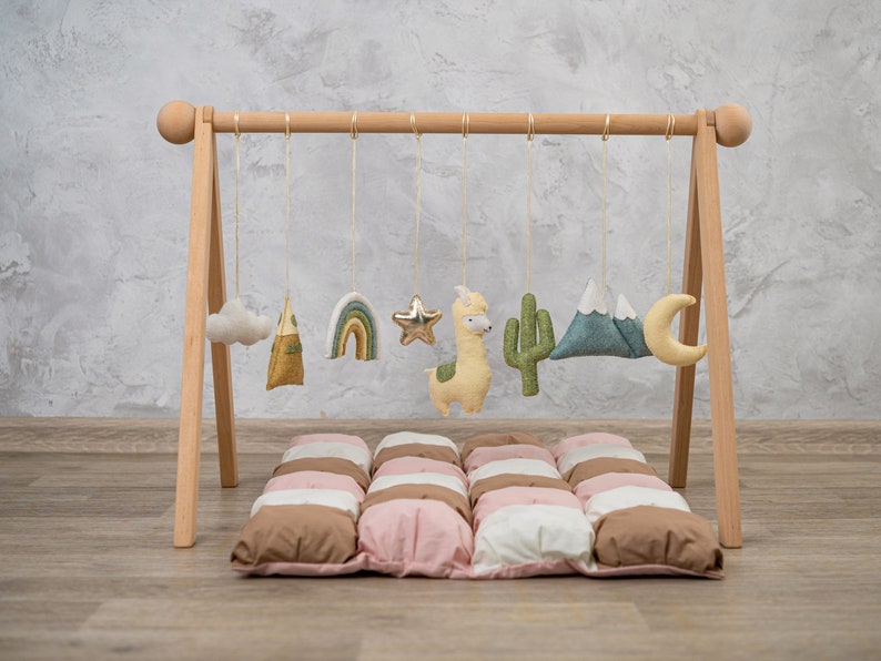 May include: A wooden play gym with a variety of hanging toys, including a cloud, a rainbow, a star, a llama, a cactus, a mountain, and a crescent moon. The play gym is set up on a brown and pink quilted play mat.