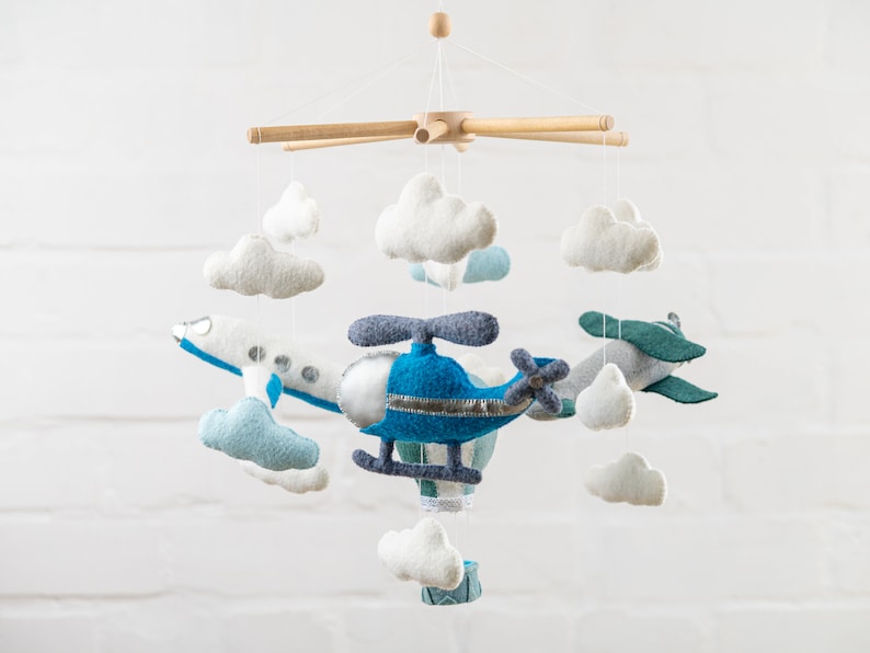 May include: A baby mobile with a blue helicopter, a white airplane, and white clouds. The mobile is made of felt and has a wooden frame.