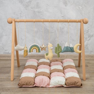 May include: Wooden play gym with hanging toys for babies. The toys include a cloud, a mountain, a rainbow, a star, a llama, a cactus, and a crescent moon. The play gym is set up on a padded play mat with brown, pink, and white squares.