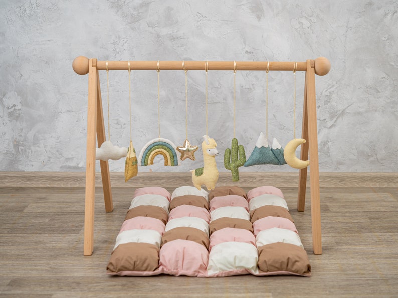 May include: Wooden play gym with hanging toys for babies. The toys include a cloud, a mountain, a rainbow, a star, a llama, a cactus, and a crescent moon. The play gym is set up on a padded play mat with brown, pink, and white squares.