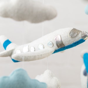 May include: A white felt airplane with blue and silver accents hangs from a mobile. The airplane has a blue stripe on the wing and silver circles on the body. The airplane is suspended from a white string.