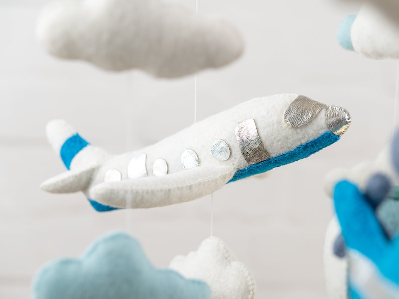 May include: A white felt airplane with blue and silver accents hangs from a mobile. The airplane has a blue stripe on the wing and silver circles on the body. The airplane is suspended from a white string.