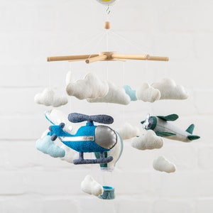May include: A wooden baby mobile with a blue helicopter, a green airplane, and white felt clouds. The mobile is hanging from a white ceiling.
