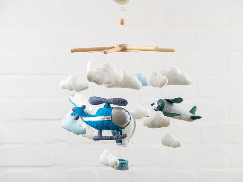 May include: A wooden baby mobile with a blue helicopter, a green airplane, and white felt clouds. The mobile is hanging from a white ceiling.