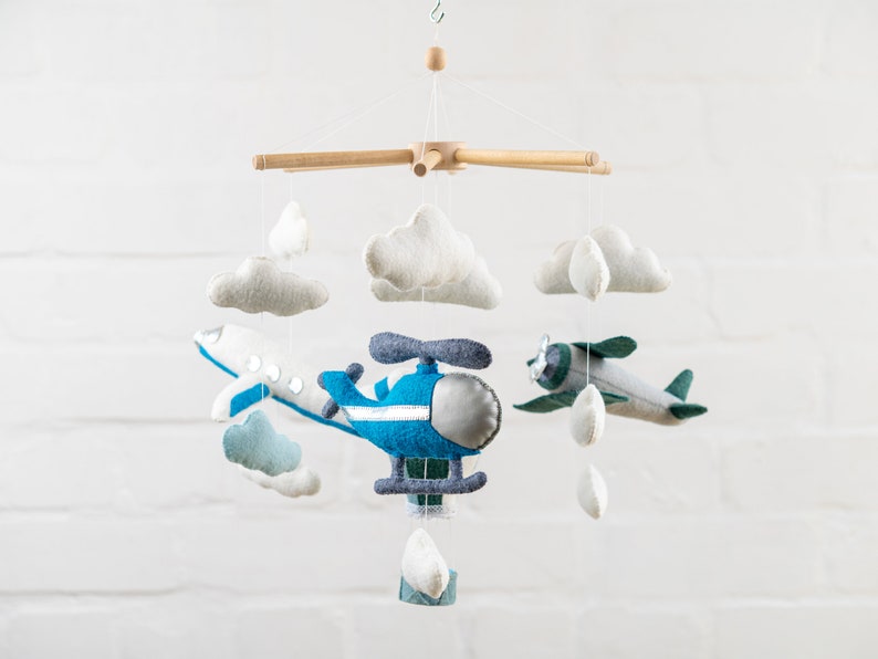May include: A wooden mobile with blue and green felt airplanes, a blue felt helicopter, and white felt clouds. The mobile is hanging from a white ceiling.